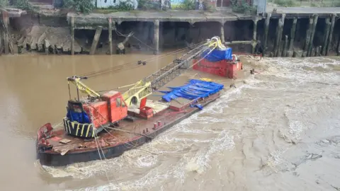 BBC/Cait Mccabe A barge with a red and yellow crane on its deck floating on a muddy river. It has sunk to the riverbed and water is lapping over the sides. There is an old wooden dock platform behind the barge on the river bank.