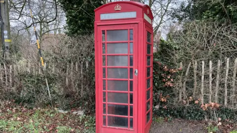 Clare Worden/BBC A traditional red phone box stands by a wooden fence with trees behind it.