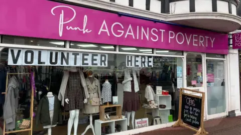 A community interest shop selling second hand goods in St Mary Church. It has a bright pink sign with the Punk Against Poverty logo on it and a small board outside that says a hundred percent of the profit from this shop goes to help local people.