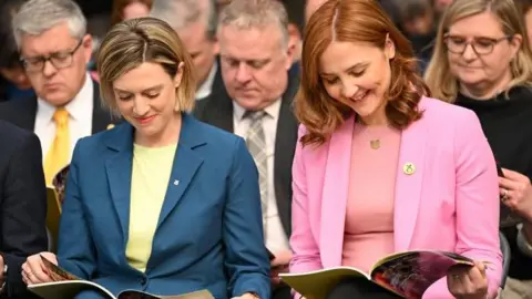 PA Media Jenny Gilruth, with a blonde bob wearing a blue blazer and yellow blouse. Sitting beside her is Màiri McAllan who has copper hair and is wearing a pink blazer and blouse.