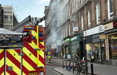 Smoke pours out of several windows and doors of a Victorian building on Union Street Glasgow in a photo taken at 1606 on Sunday 8 March. There is a fire appliance in the foreground and three firefighters can bee seen in the street.