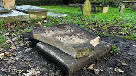 A broken gravestone, two large pieces of stone, one on the ground with mud and leaves around it, with the other piece of stone on top of the other.