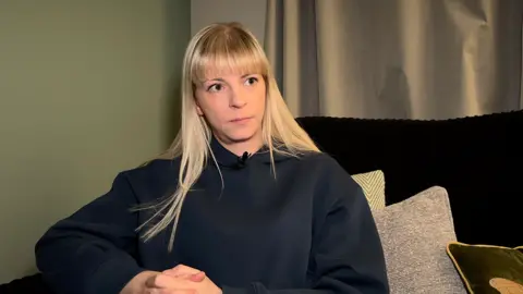 BBC A woman with blonde hair is wearing a black top and looking off to the right hand side of the shot. She is sitting on a sofa with a number of cushions.