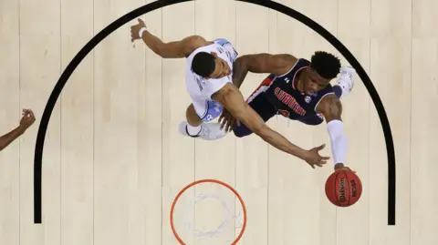 Getty Images Malik Dunbar attempts to score a lay-up while playing for the Auburn Tigers. The camera view is above the basket, looking head down at Dunbar, in blue, and his opponent, in white, who is trying to stop him. 