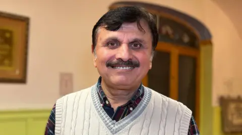 Sarfaraz is wearing a light-coloured knitted sleeveless sweater over a checked shirt stands indoors in a room with green panelled walls. A framed artwork hangs on the wall behind, and a wooden doorway is visible to the right.