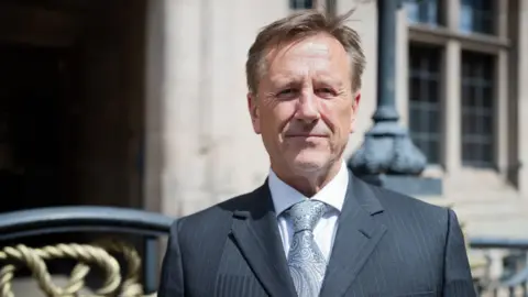 Staffordshire County Council Martin Murray is standing in front of the county council buildings in Stafford looking at the camera, he is wearing a grey tie, shite shirt and black suit jacket.