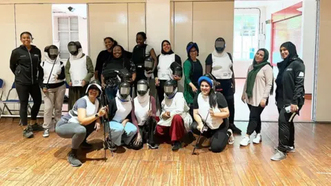 Home Girls Unite A group of women, some in fencing gear, stand in a room with a wooden floor