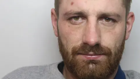 Krzysztof Orzechowski's mugshot. He has a brown beard and short hair. He is wearing a grey prison-issue jumper and a blue prison-issue T-shirt.