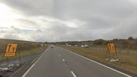 A view of a road with two lanes on each side, separated by a grass verge. There are yellow road signs on the left and right showing an upcoming lane is closing