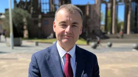 Gary Ridley has short dark hair. He wears a red tie, blue suit jacket and white shirt.