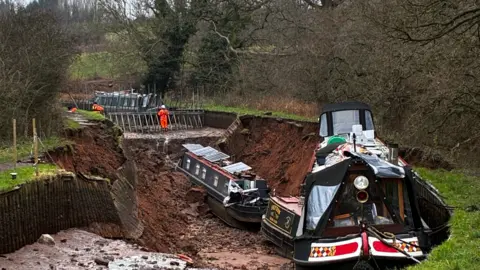 Two boats in a dry hole in a canal. Another boat hangs at the edge of the hole. On the other side is a man dressed in high-visibility orange gear, leaning against metal spikes installed in the dry canal bed.