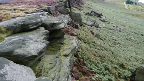 Save Walshaw Moor campaign group The image shows a moorland escarpment with large, weathered gritstone rocks forming a cliff edge. Below the rocks the hillside drops into a wide expanse of open moor covered in heather, grasses, and patches of green vegetation. 