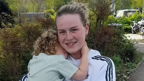 Charlie Flynn has blond hair tied back and is wearing a white Addidas hooded top. Her daughter, who has curly blond hair, is in her arms and is hugging into her. 