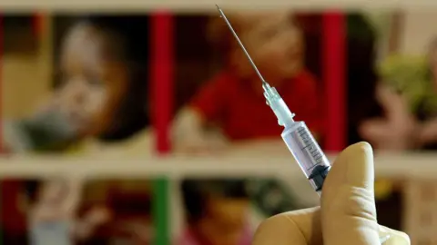 Library image of a clear, colourless syringe photographed in sharp focus. In contrast, young children in the background of the shot are in very soft, hazy focus