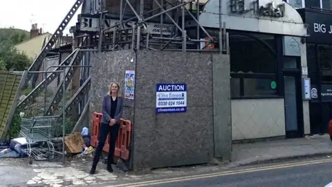 Laura Kyrke-Smith Labour MP Laura Kyrke-Smith next to the scaffolding in Aylesbury. She is tall and has blonde shoulder-length hair. There is a shopping trolley and rubbish next to her and on the other side of the scaffolding there is an Indian restaurant.