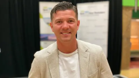 Head and shoulders shot of Luke Campbell. He is wearing a light-coloured jacket and is smiling at the camera. There is a display board in the background.