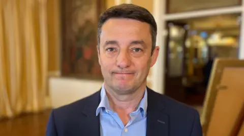 BBC Boley Smillie - a man wearing a navy suit and light blue shirt, standing in a large hall room which is blurred.