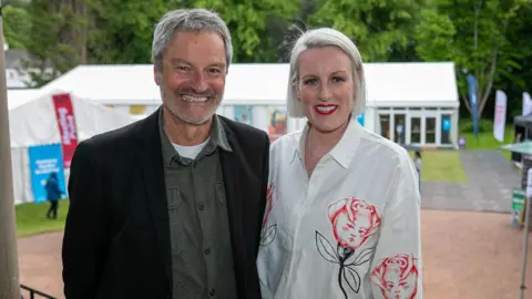  Gavin Esler and Steph McGovern - he has grey hair and grey stubble and is wearing a dark jacket with a green shirt underneath. She has blonde hair and is wearing a white blouse with a rose pattern on it.