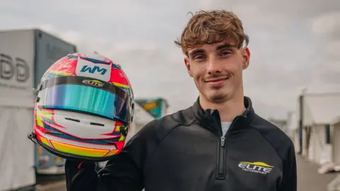William Macintyre, a young man who is standing outside holding a racing helmet. He is looking directly at the camera and smiling and is wearing a black zipped-up jacket.