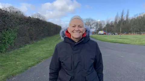 A woman with short grey hair is wearing a black hooded, padded coat. She is standing in a park and smiling