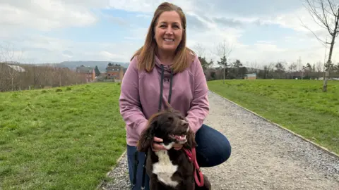 Viv, a woman with long hair, wears a dusky pink hoodie and dark blue jeans. She is crouching down on a gravelled pathway that runs through a grassed area, with houses in the distance. Her hands are resting on Dolly, a brown cockapoo with a white chest, who is sitting in front of her