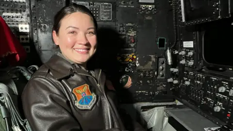 Captain Christina Herman sat at the control panel of a bomber plane. She is smiling at the camera and wearing a leather bomber jacket with the United States Air Force emblem on it.