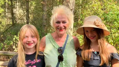 A woman and two girls standing together on a woodland path, with trees and a wooden fence in the background.