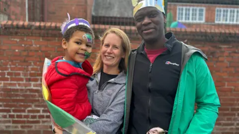 Elspeth and John Thompson, john in bright green jacket and black vest, elspeth holding son in red jacket, wearing a grey hoody, pictured against red brick wall