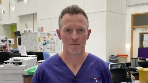 Dr Courtney is looking at the camera. He is wearing dark blue scrubs and has short greying hair and stubble.