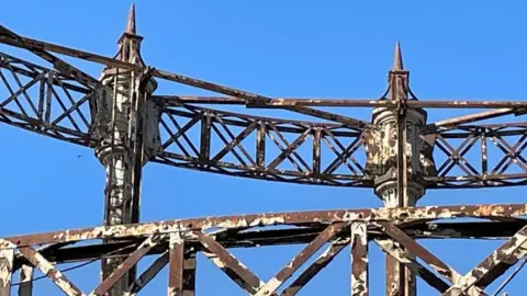 Andrew Turner/BBC The Great Yarmouth gasometer stands in a blue sky, with it's peeling paint and rusting of the columns and lattice work. With finials on the top it is is highly decorated, with it being awarded Grade II listed status.