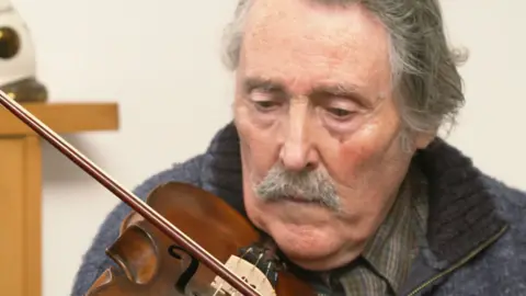 BBC Man playing fiddle, he has longish grey hair and a moustache. He is wearing a checked shirt under a blue jacket.
