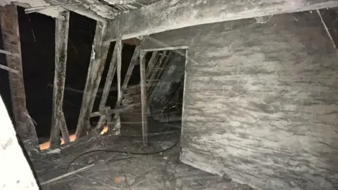 Karina Rose/Woodbridge Red Lion A dark room with ancient beams on the left, a doorway and a grey wall