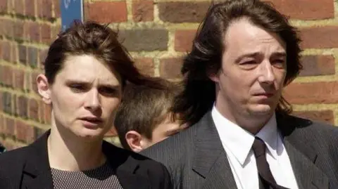 A woman with brown hair, wearing a black suit and dotted black shirt, is walking next to a man with brown hair. He is wearing a dark grey suit, white shirt and dark grey tie.