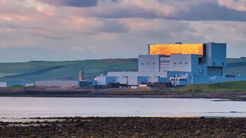 Torness nuclear power station with the sun reflecting on the windows. It is situated behind a stretch of water with rockery in the foreground.