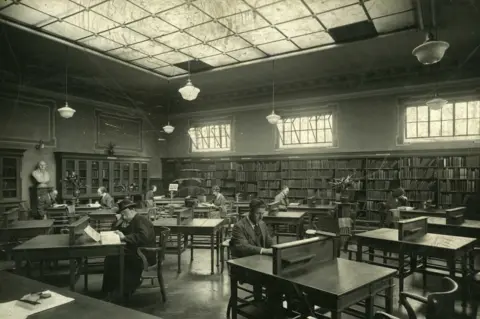 Gateshead Library Several men sitting at desks in the library's reading room. A large glass ceiling is above them allowing light to enter the building. Books are shelved along the walls.
