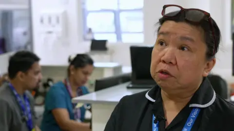 Milady Doinog with shoudler length hair in a pony tail wearing brown-rimmed glasses and a black NHS medical uniform and blue NHS landyards around her neck