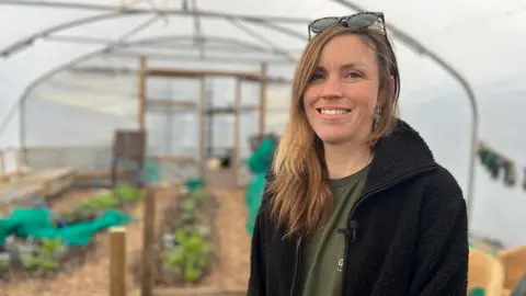 A women is looking at the camera and smiling. She has a pair of sunglasses on her head. She is wearing a khaki green top with a black zip-up fleece over the top. She has brown hair with a nose ring and silver hanging earings. 
She is standing in a polytunnel and there are plants behind her. 