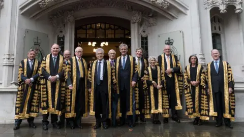 Getty Images The judges of the Supreme Court gathered outside the doors. There are 10 men and two women standing in a row wearing the black and gold robes of the court.