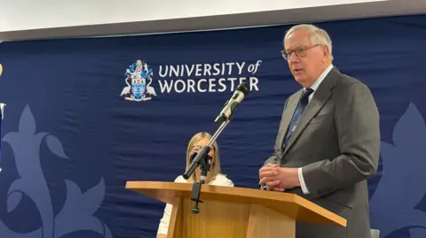 A man wearing glasses in a shirt, blue tie and grey jacket is standing at a lectern. A blue background is behind him, along with a woman with blonde hair whose face is partially obscured by a microphone stand on the lectern.