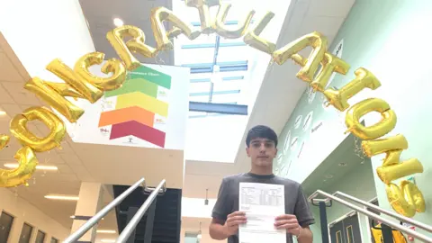 Mick Lunney/BBC A teenage boy stands at the bottom of a staircase holding a results sheet, with a large gold “Congratulations” balloons overhead.