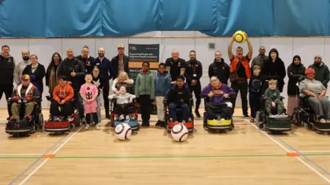 Rotherham United Community Trust's powerchair football team - an accessible form of the sport for wheelchair users. Coaches, parents and players of all ages - both children and adults - are stood in a group.