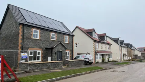 A row of new build homes, all of them with solar panels on the roof. One of the houses is in dark brick, a few are rendered in white plaster and another is in a reddish brick. The homes are empty and there are some signs of construction work such as a small van and metal fencing. 