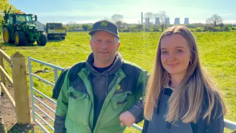 A man and women stand by a metal gate attached to wooden fencing. There is a large green field behind them with a tractor and truck parked in it. The woman has long fair hair and a grey fleece. The man is wearing a black cap, green overalls and a grey hoodie. There are trees and industrial chimneys in the distance.