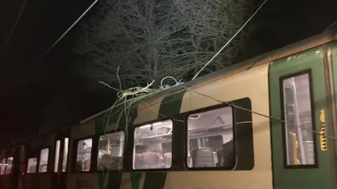 A green and cream train pictured at night, with wires tangled on its roof. The train lights are on and a passenger can just be seen sitting inside.