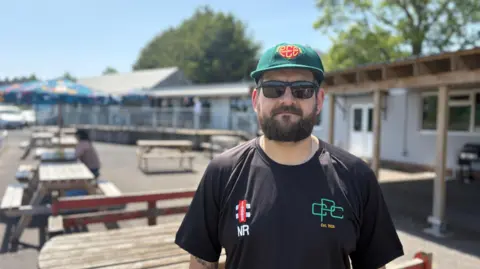 Nathan Redwood stands in front of the clubhouse wearing a black Purnell Cricket Club t-shirt, featuring his initials and the club logo. He has a dark beard and is wearing sunglasses and a green club cap. 