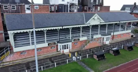 Shaun Whitmore/BBC A Victorian era football stand with pastel green paint and tiled roof can be seen, with houses behind it and grass to the front.