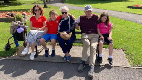 Eiry Miles A family sat on a bench in a park, with a walker parked next to them. From left to right: A woman with brown hair and sunglasses, in a red top and grey striped skirt, next to a young boy in a Wales football shirt and blue shorts, who has his arms around a woman with short grey hair dressed all in dark blue with a white scarf and brown handbag. Next to her is an older man in a grey cap, purple sweater vest and beige trousers. He has his arm around a young girl with brown hair in a pink top