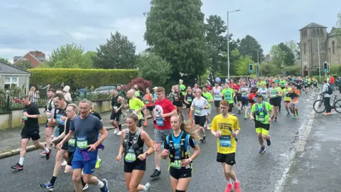 PA Numerous people in various coloured sportswear running along a road. There are people watching on from the footpath. There is a stone house and building in the background along with a number of green trees and hedges.