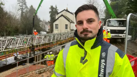 Man in a bright yellow high-visibility jacket stands at a roadworks site where a temporary metal bridge is being installed. Behind, several workers in orange safety gear are visible on and beneath the structure, with a green concrete pump extending from a white lorry. A white house and trees can be seen in the background under grey skies