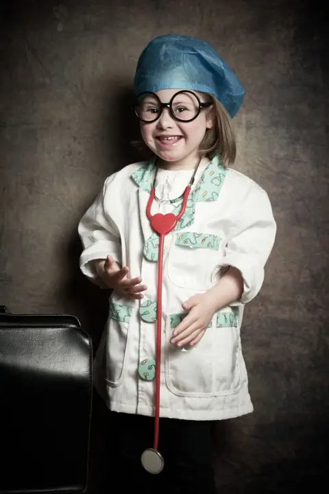 Debbie Todd Layla, a young girl with shoulder length brown hair wearing a blue surgical hat, large round black-framed glasses, a child's nurse coat and large red stethoscope around her neck, smiles at the camera